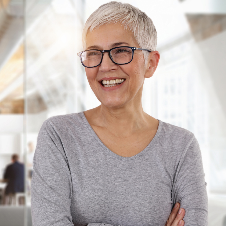 Smiling older woman with short gray hair and glasses stands with arms crossed in a modern, bright office setting.