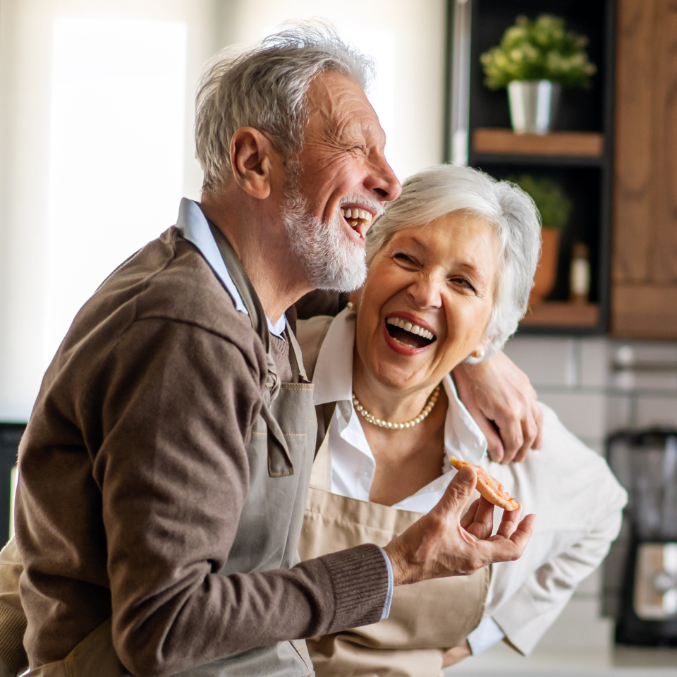 Two older adults with gray hair, wearing aprons, smile and laugh together in a bright kitchen. One holds a cookie; they appear to be enjoying each other's company.