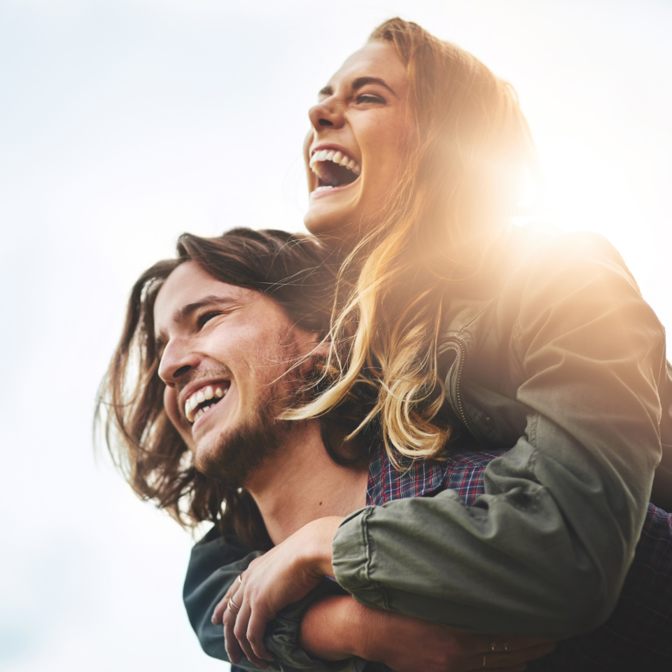 A woman laughing while giving a piggyback ride to a smiling man outdoors, with sunlight shining behind them.