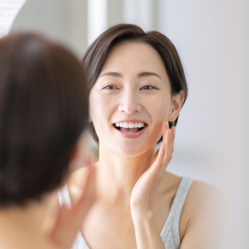 A woman smiles while looking at herself in a mirror, touching her face with one hand.