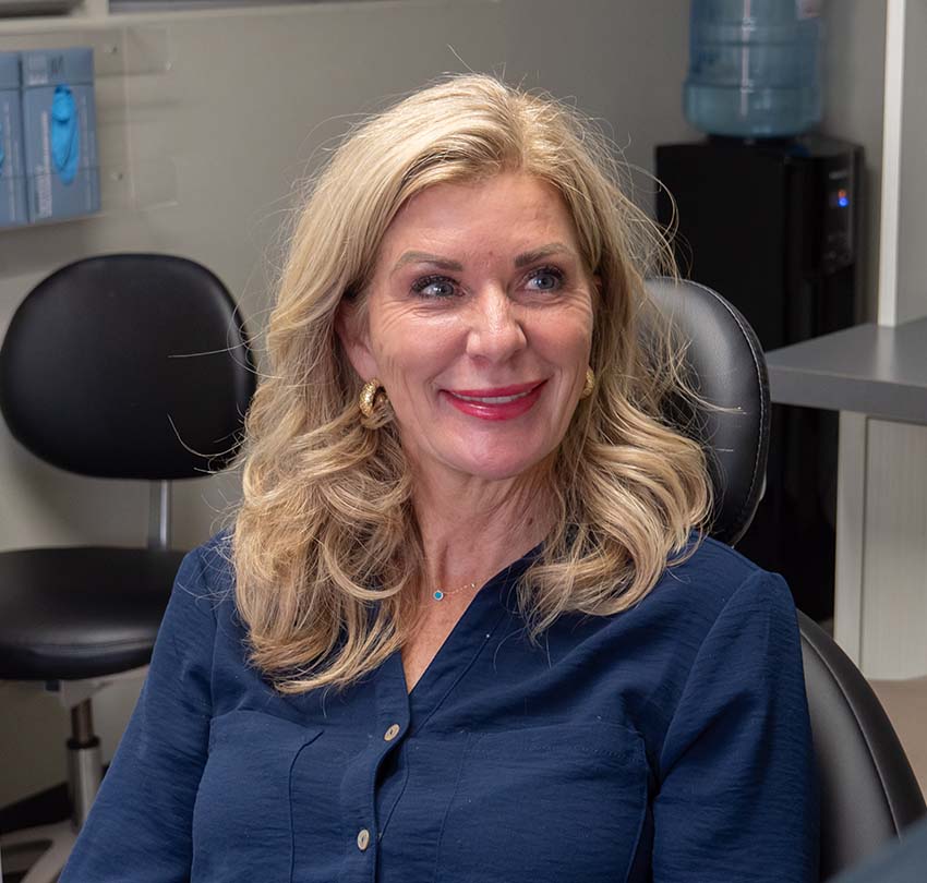 A woman with long blonde hair and a navy blouse sits in an office chair, smiling. There is a black chair, a table, and a water cooler in the background.