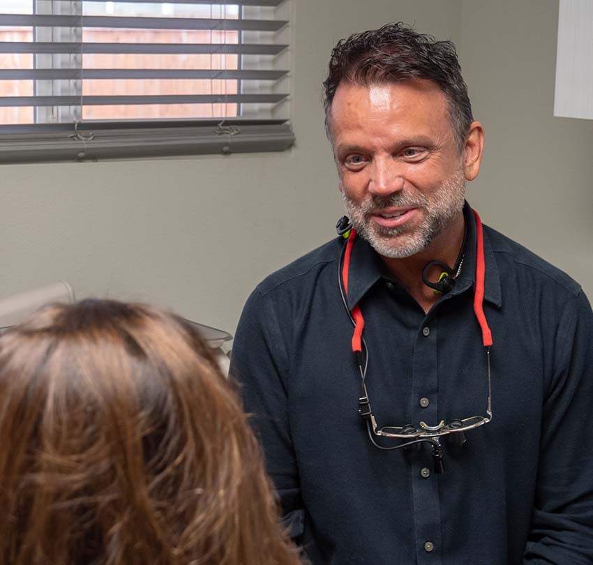 A man wearing a black shirt and eyeglasses around his neck talks to a seated woman in a medical or office setting.
