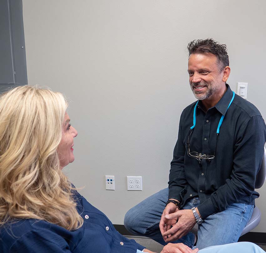 A man in a dental office talks with a woman sitting in a chair. Both appear to be having a friendly conversation.