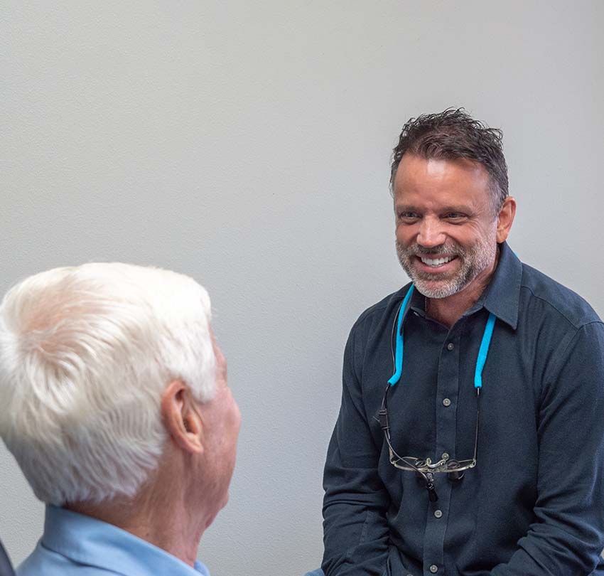 A man with a beard and glasses around his neck smiles while talking to an older man with white hair in a light blue shirt in a plain room.