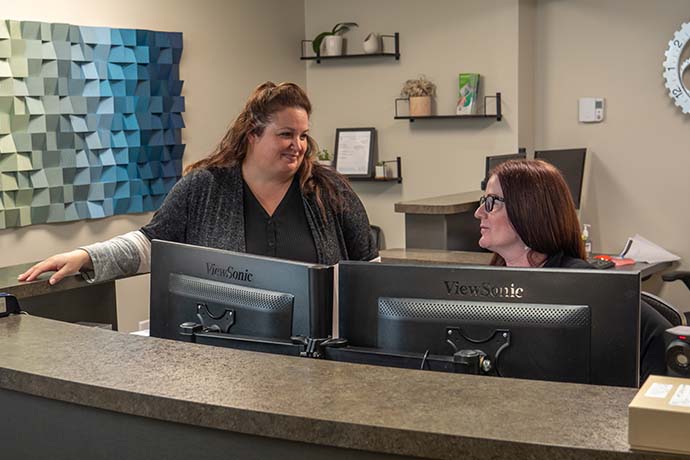 Two women talk at an office reception desk. Both are standing behind computer monitors, with office decor and supplies visible in the background.