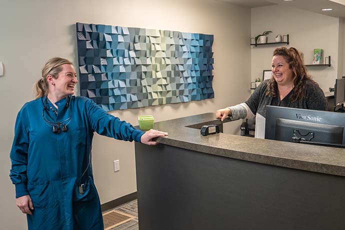 A woman in scrubs stands and smiles while talking to a woman behind a reception desk in an office setting.