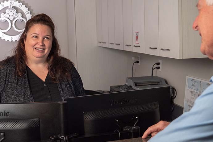 A woman stands behind a reception desk, smiling and speaking with a man across the counter in an office setting.