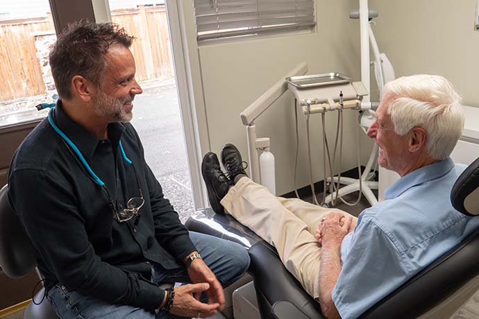 A dentist talks with an older male patient seated in a dental chair inside a dental office, with dental tools visible in the background.