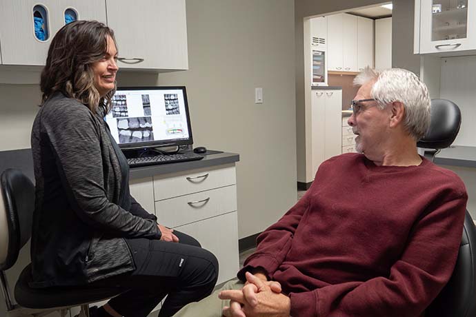 A woman and a man are seated in a dental office, conversing. Dental X-rays are displayed on a computer screen in the background.