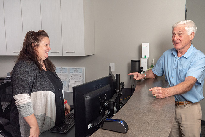 An older man speaks with a female receptionist at a front desk in an office setting.