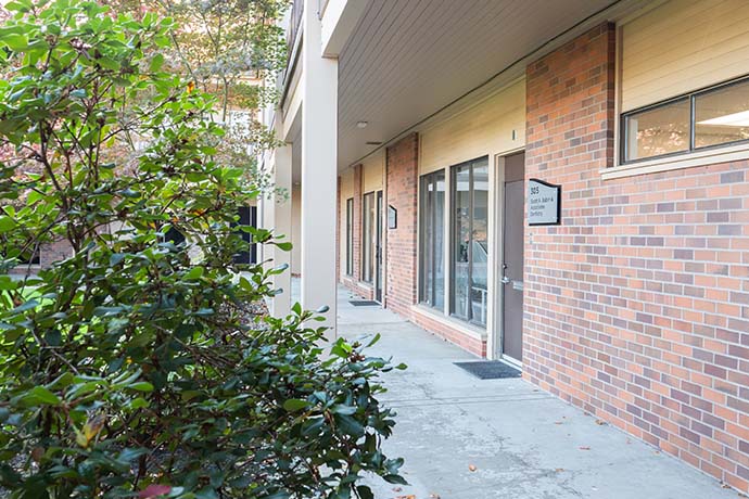 A brick building with large windows and a sign reading "35" beside the entrance, partially obscured by green bushes along a concrete walkway.