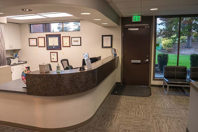 Reception area with a curved desk, office chair, certificates on the wall, exit door, waiting chairs, and large window showing greenery outside.