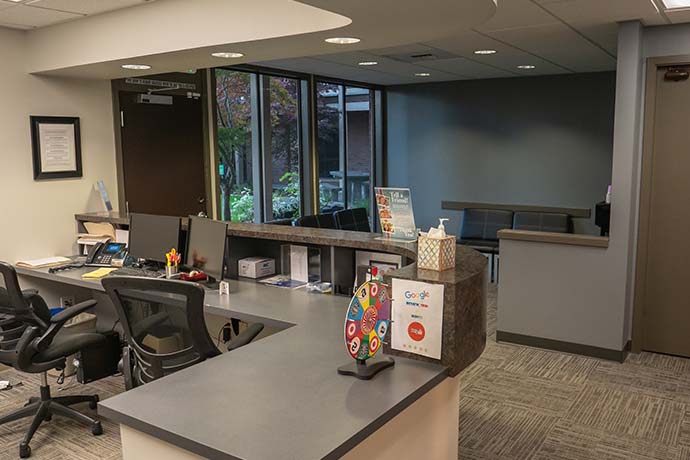 Reception area with front desk, office chairs, computers, and a waiting area with gray seating; large windows show greenery outside. A colorful prize wheel and tissue box are on the counter.