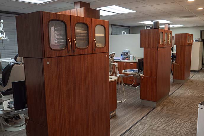 Row of dental exam cubicles with wood-paneled cabinets and dental equipment, under fluorescent ceiling lights in a clinical office setting.