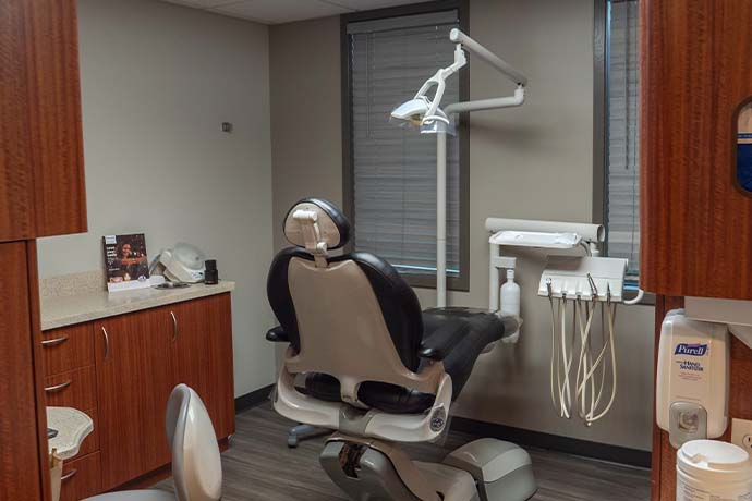 Dental exam room with patient chair, overhead light, dental tools, cabinets, and a window with blinds. Hand sanitizer is mounted on the wall near the entrance.