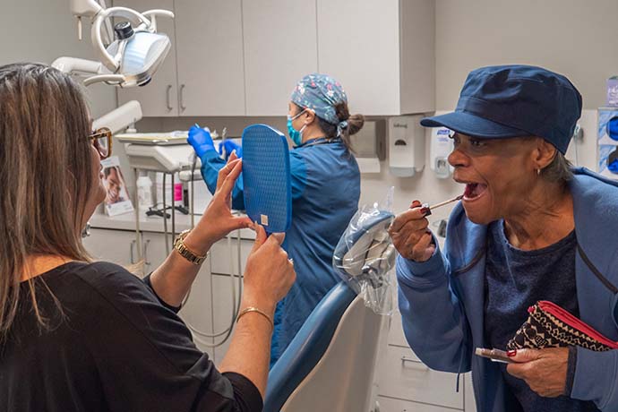 A woman looks into a mirror held by a dental professional while applying lipstick; another dental staff member works in the background.