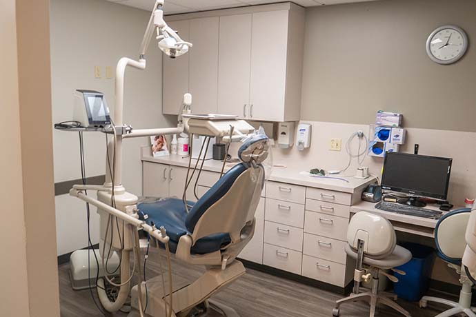 A dental office with an empty exam chair, overhead light, computer, cabinets, and medical supplies on the countertops.