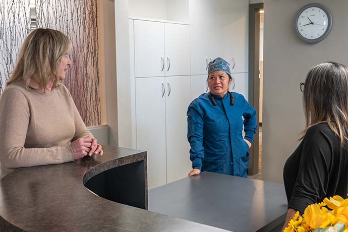 Three women talk at a modern reception desk in a medical or dental office. One wears medical scrubs, while the others are in casual clothing. A wall clock shows the time as 11:10.