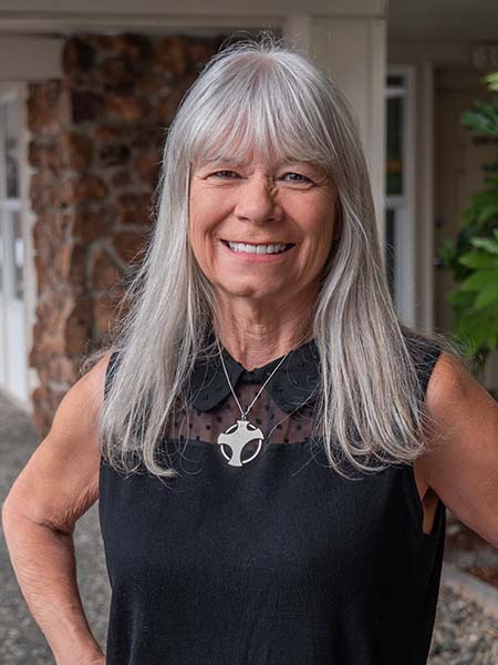 Older woman with long gray hair wearing a sleeveless black top and silver pendant, standing outdoors and smiling at the camera.