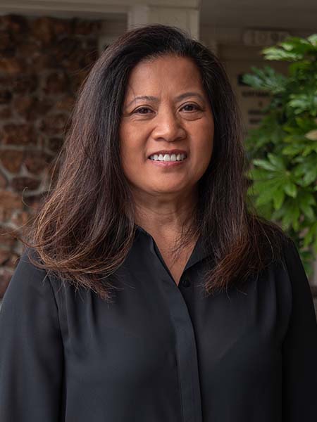 Woman with long dark hair wearing a black blouse stands outdoors, smiling at the camera. A stone wall and green plant are visible in the background.