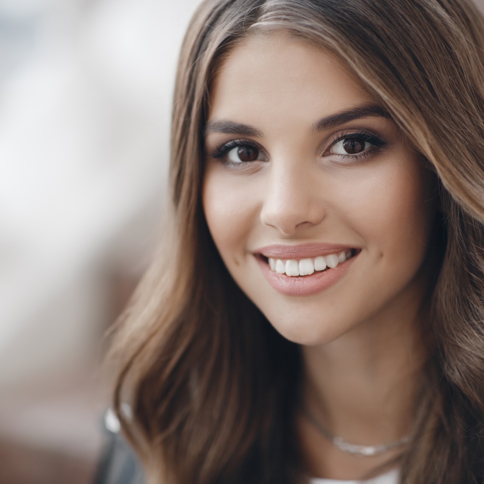 A young beautiful woman with long, hair and brown eyes, beautiful makeup, pink plump lips, sweet smile,brunette, spending her time outdoors in the summer in the city, dressed in a white t-shirt , posing in the sunlight. Portrait of happy young woman
