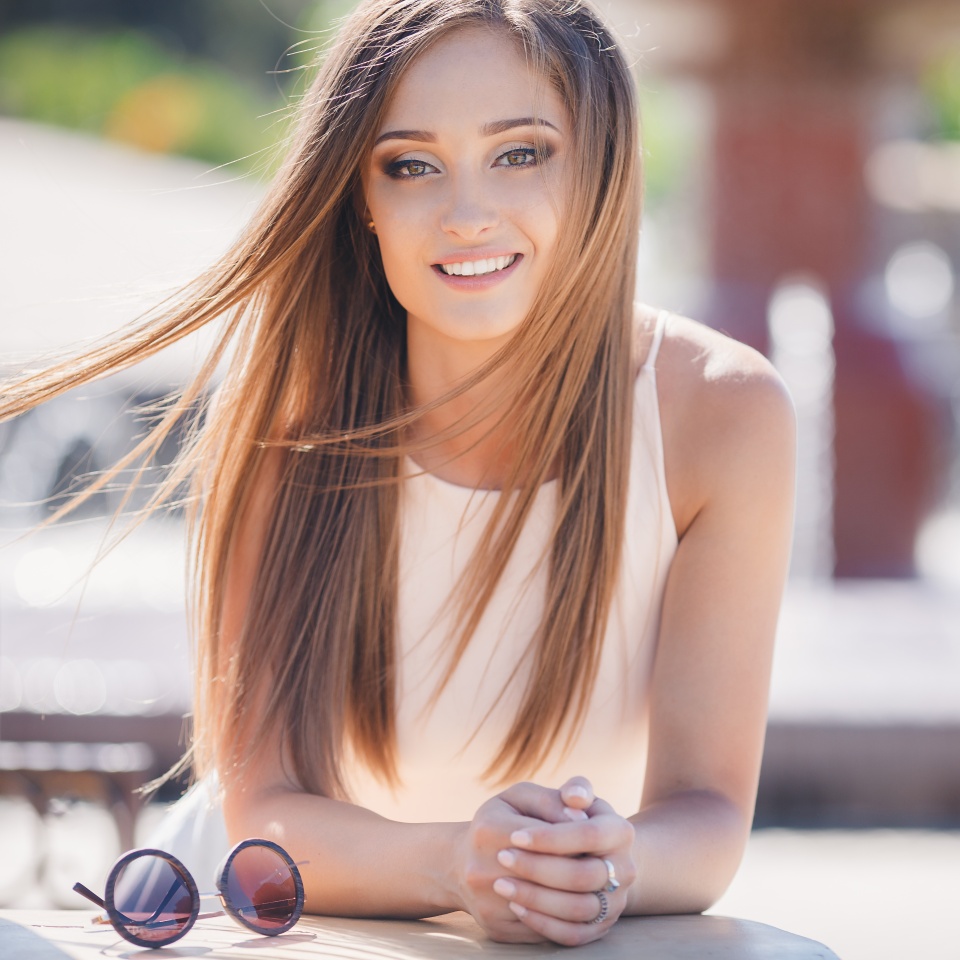 Young,beautiful,attractive woman,a brunette with long straight hair and gray eyes,a beautiful smile and straight white teeth,dressed in a white shirt,sitting at a table in a city cafe,posing against a green urban landscape.