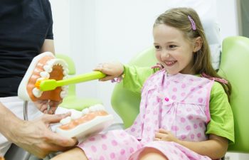 Smiling little girl in dentists chair being educated about proper tooth-brushing by her paediatric dentist. Early prevention raising awareness oral hygiene demonstration concept.