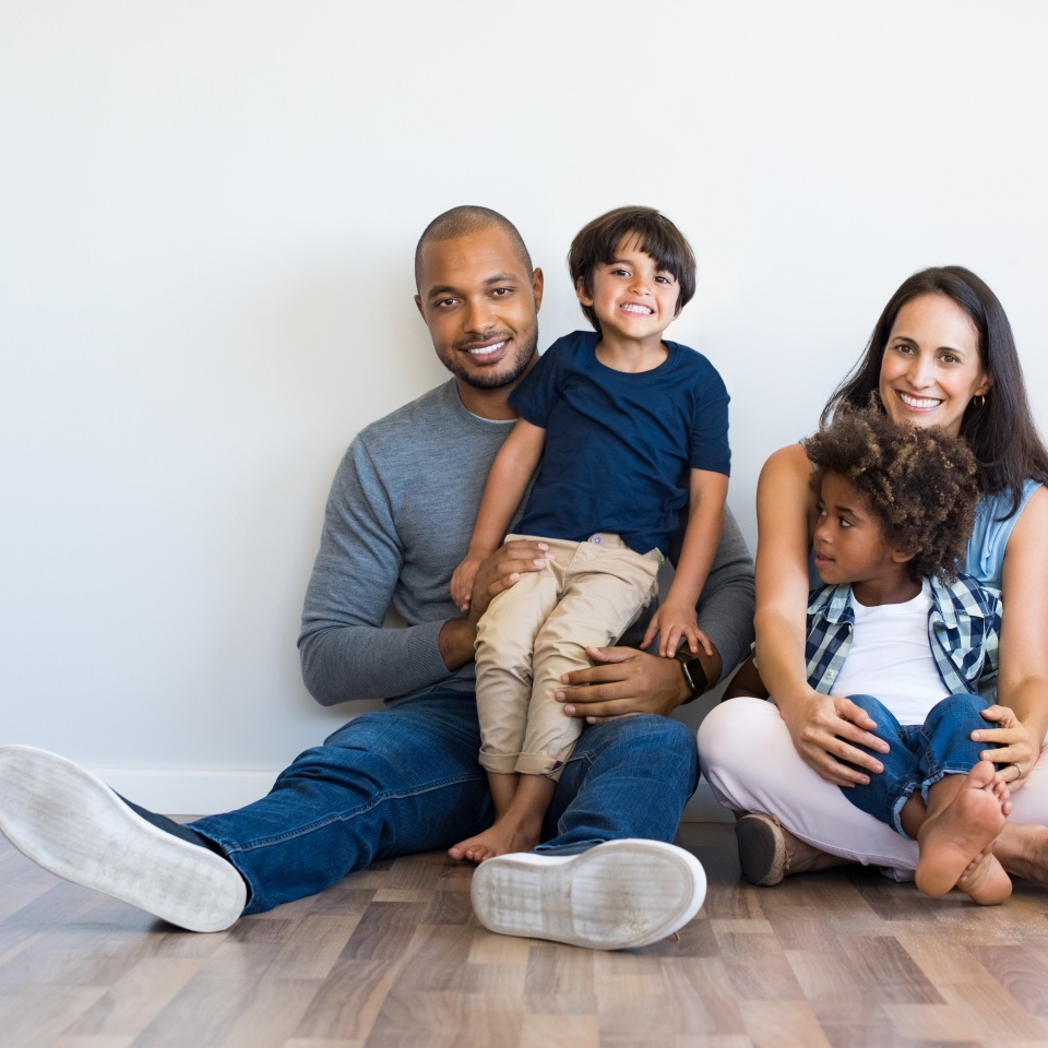 Happy multiethnic family sitting on floor with children. Smiling couple sitting with two sons and looking at camera. Mother and black father with their children leaning on wall with copy space.