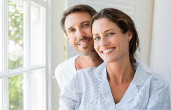 Happy young couple sitting near window in the bedroom. Portrait of smiling man and woman looking at camera at home. Couple in pajamas during the morning.