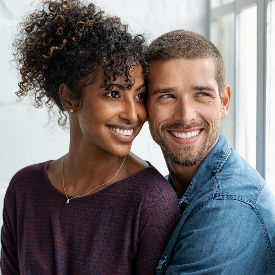 Loving multiethnic couple embracing and sitting near window. Happy girlfriend and smiling boyfriend looking away and thinking about their future together. Young man and african woman contemplating.