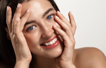 Radiant and glowing facial skin, women cosmetics. Beautiful girl model touching her nourished, hydrated clean face and smiling happy, blue eyes and healthy skin, white background