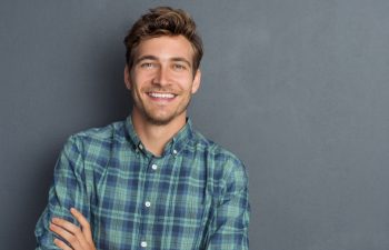 Young handsome man leaning against grey wall with arms crossed. Cheerful man laughing and looking at camera with a big grin. Portrait of a happy young man standing over grey background.