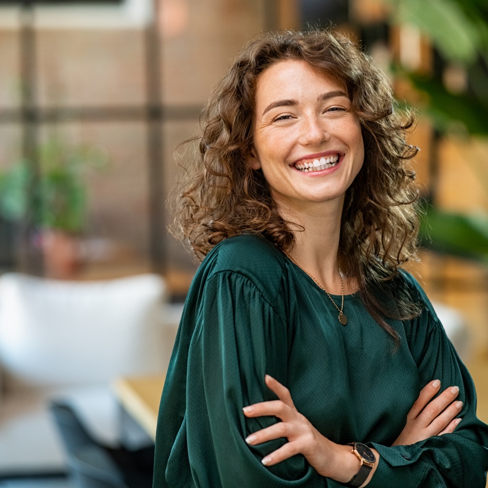 Portrait of young smiling woman looking at camera with crossed arms. Happy girl standing in creative office. Successful businesswoman standing in office with copy space.