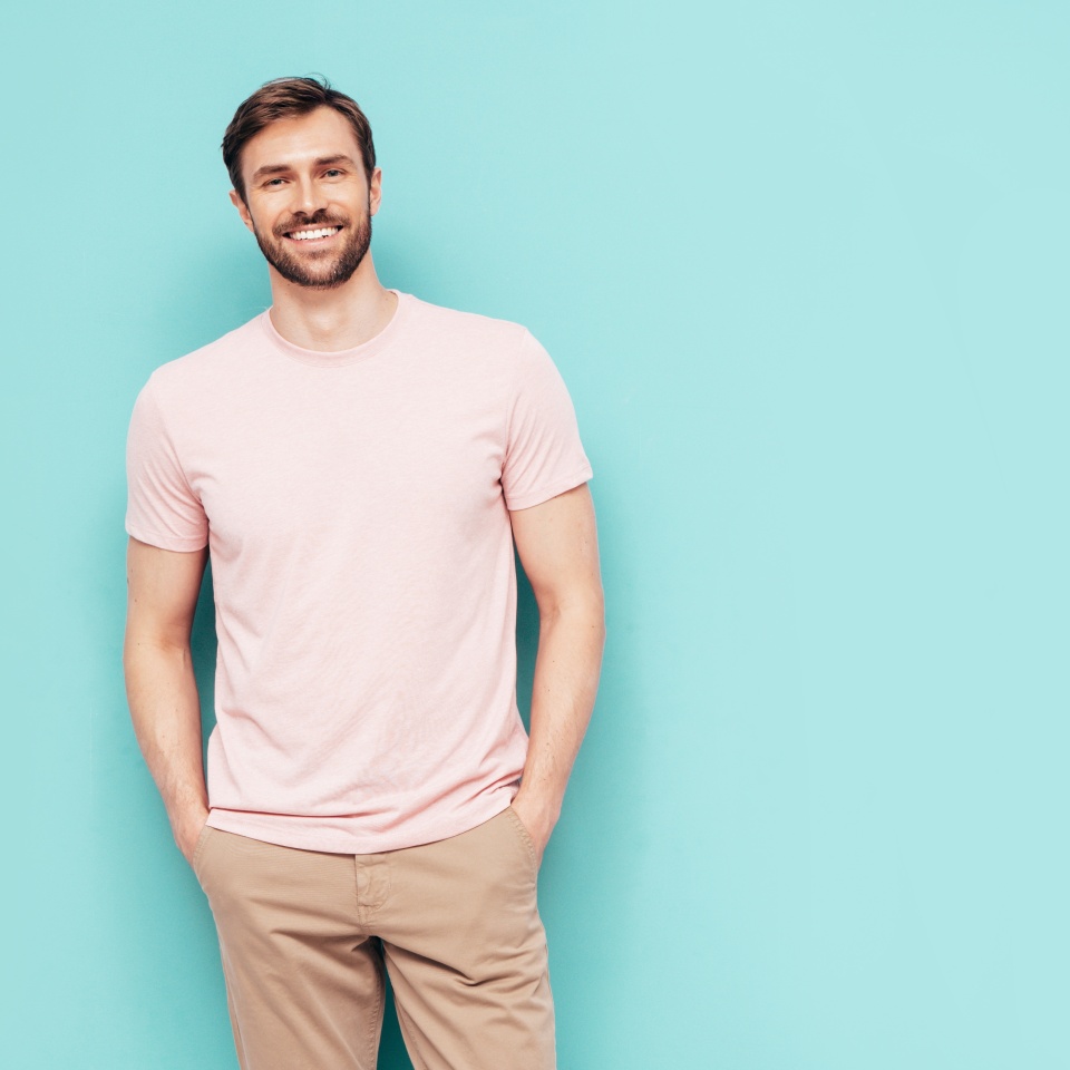 Portrait of handsome smiling stylish hipster lambersexual model. Sexy man dressed in pink T-shirt and trousers. Fashion male isolated on blue wall in studio
