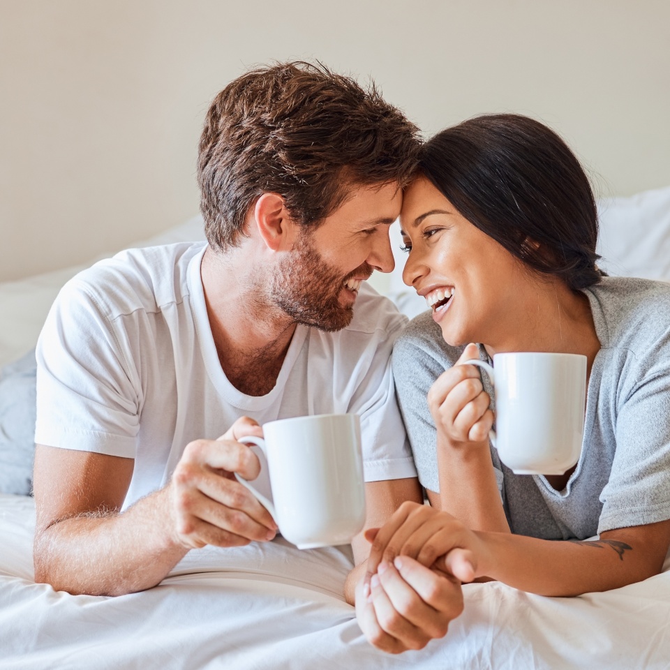 Love, happy and a couple with coffee in bed on beautiful morning at home. Weekend, wake up and smile, woman and man relax with hot drink in bedroom. Romance, happiness and drinking sweet tea together