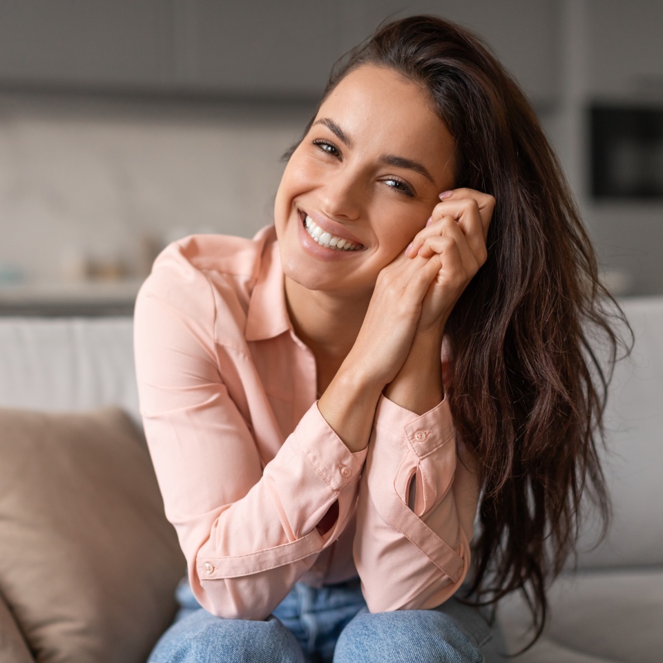 Radiant woman with contagious smile poses playfully, hands gently cradling her face, capturing moment of genuine happiness and effortless charm