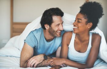 Happy, carefree and laughing couple having fun lying in bed together. Interracial husband and wife bonding and showing affection while talking. Smiling lovers enjoying time indoors being playful.
